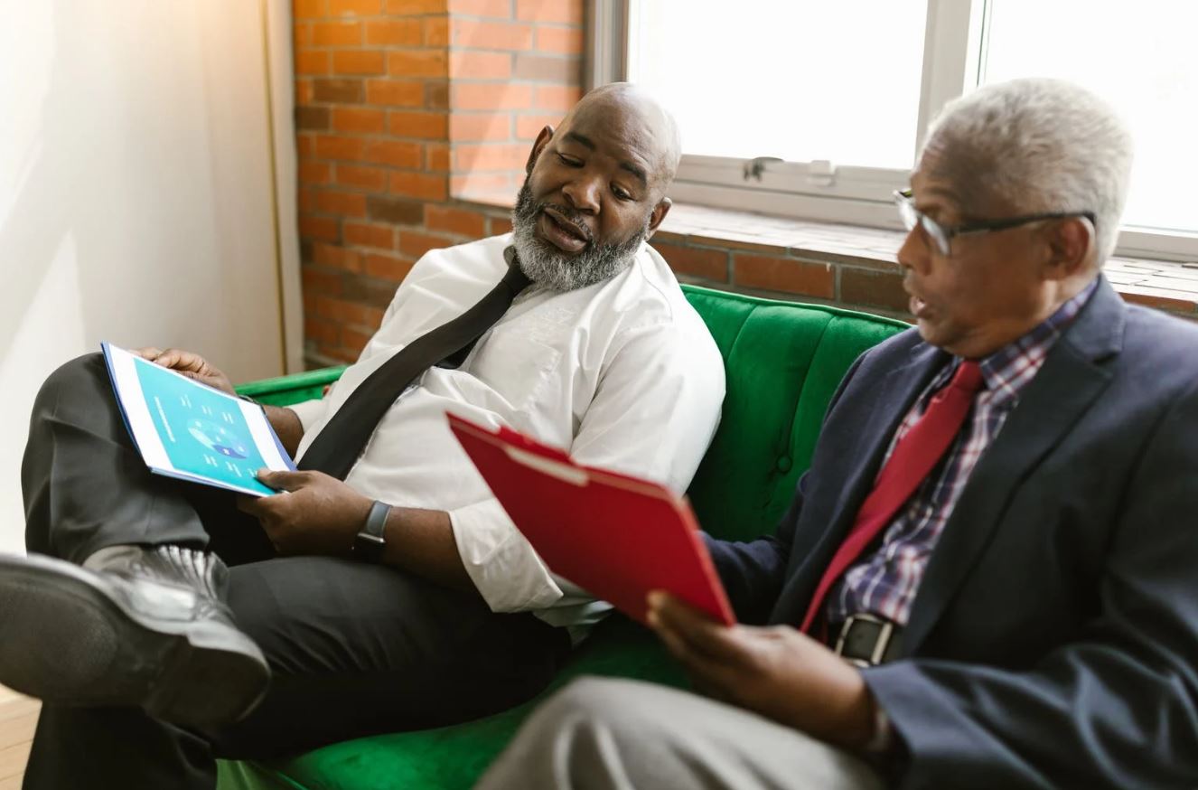 Two men sitting on a green couch, looking at open folders and talking in a brick-walled room.