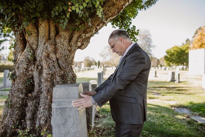 Man in a dark suit standing beside a gravestone under a large tree in a cemetery.