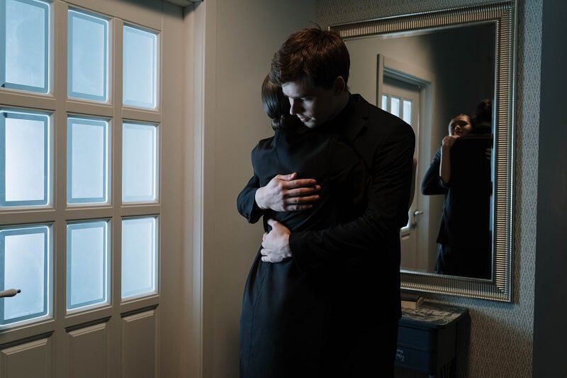 Two people embracing in a dim hallway beside a window and mirror, one holding a hand to the other's back.
