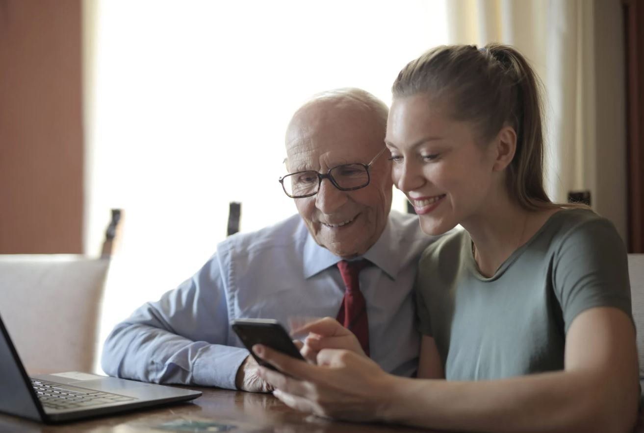 Two people smiling at a smartphone beside a laptop in a bright room