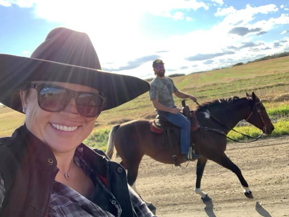 Smiling woman in a black hat and sunglasses on horseback, with another rider on a trail under a bright sky.