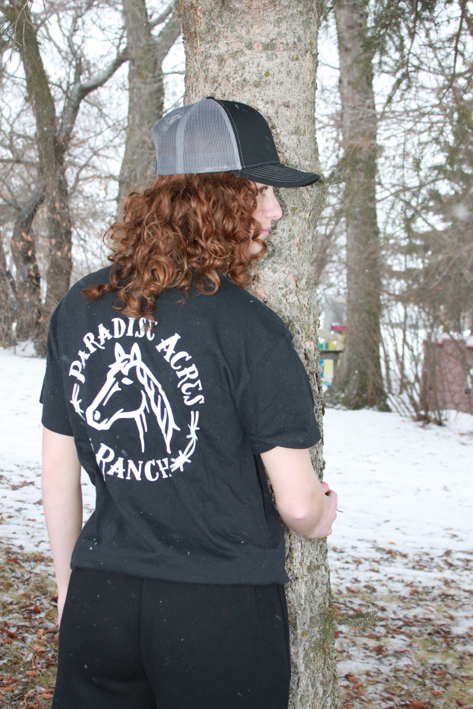 Person with curly hair and a cap in a black shirt, standing outdoors in a snowy wooded area.