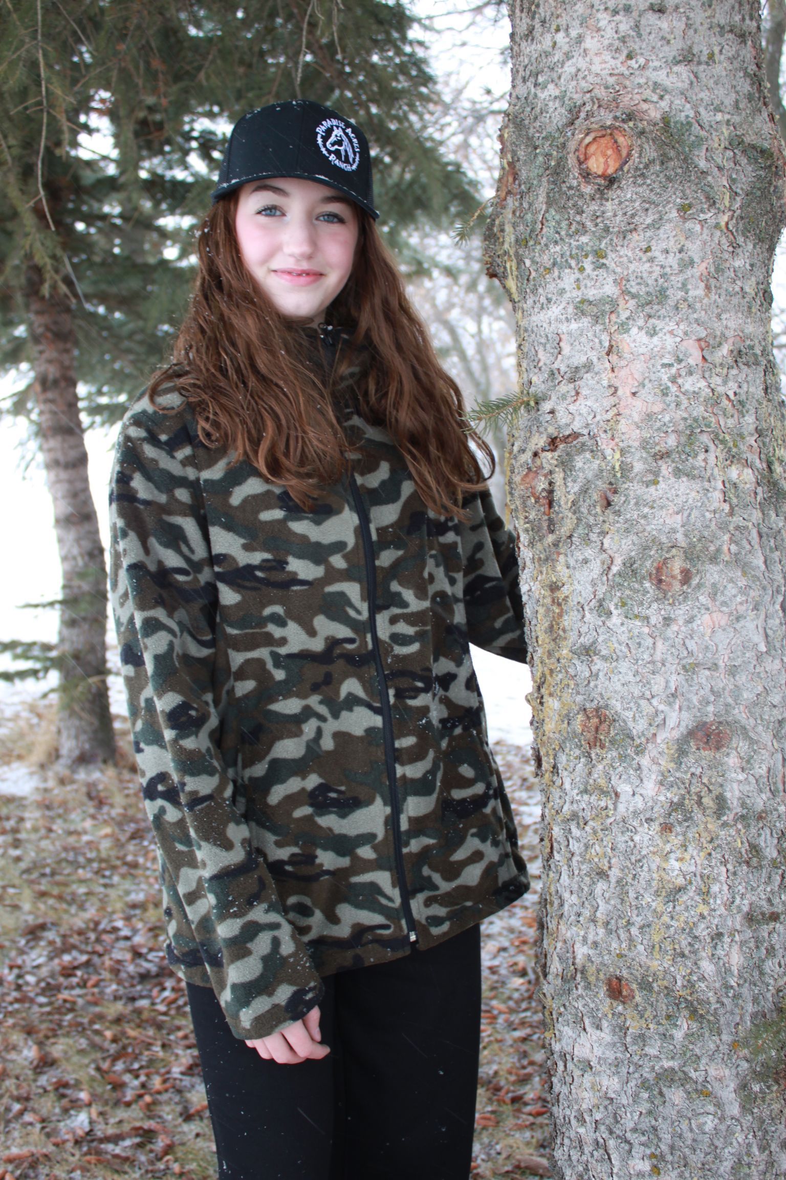 Woman in camouflage jacket and black hat standing beside a tree in a wooded area.