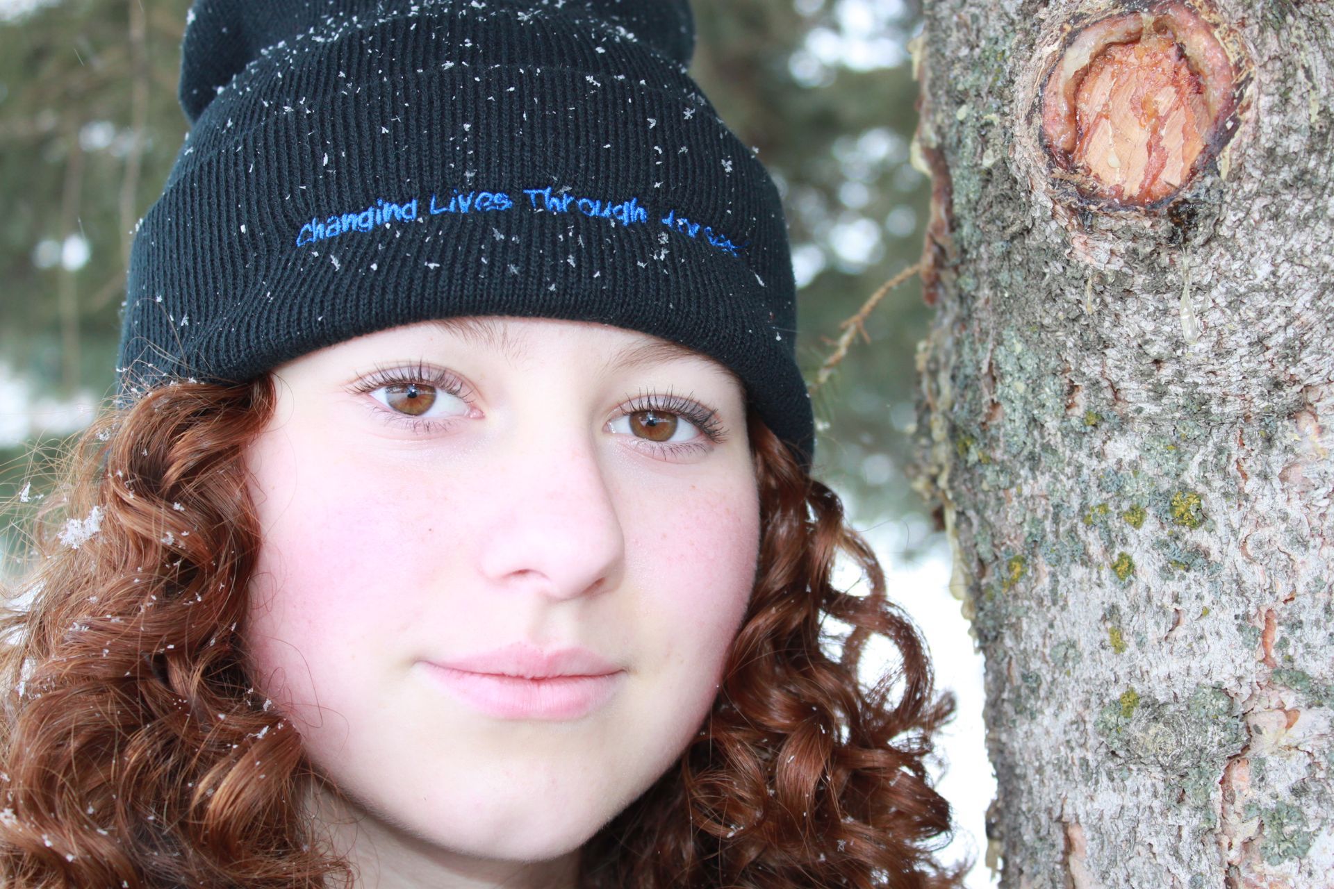 Close-up of a person in a black beanie beside a tree trunk outdoors in winter.