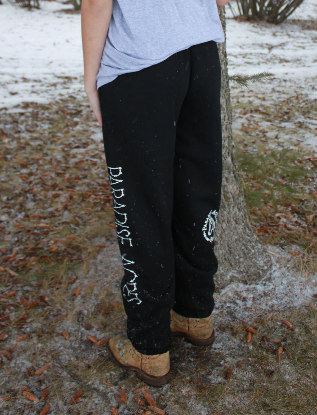 Person standing outdoors beside a tree, wearing a blue shirt, black pants, and brown boots.