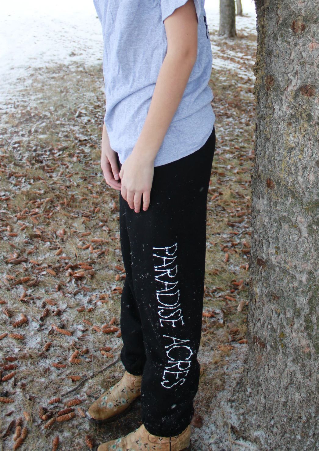 Person standing outdoors beside a tree, wearing a blue shirt, black pants, and brown boots.
