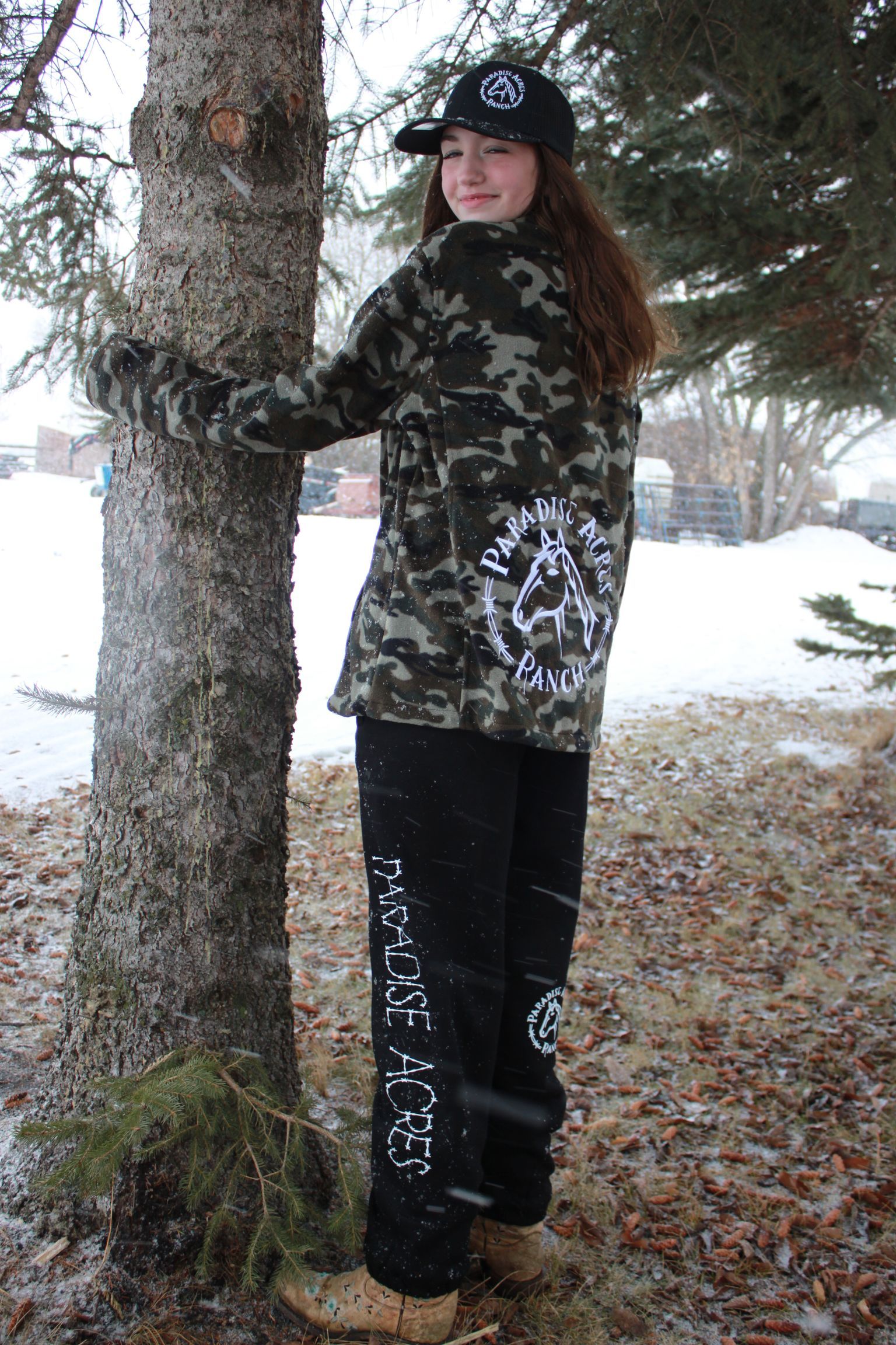 Woman in camouflage jacket and black hat standing beside a tree in a wooded area.
