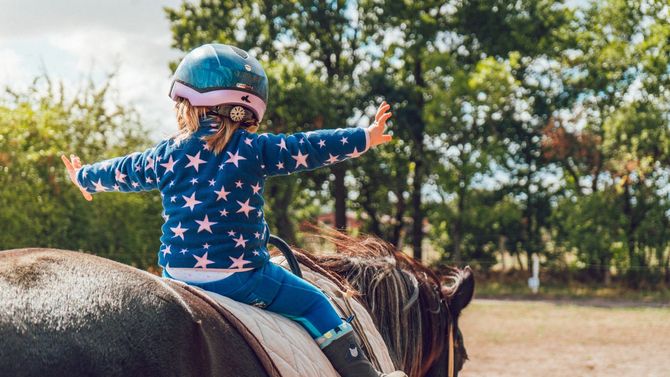 Child in blue star-patterned outfit and helmet riding a horse with arms outstretched outdoors.