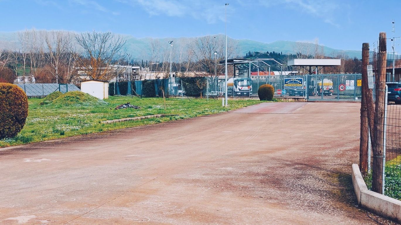 Strada sterrata che conduce a una zona industriale recintata, con montagne in lontananza e cielo azzurro.