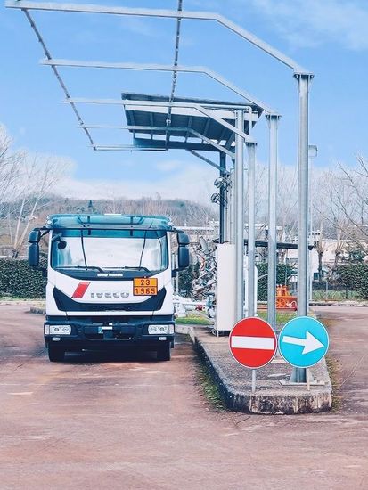 Camion di carburante sotto una tettoia metallica in una stazione di servizio con segnaletica stradale; cielo azzurro sullo sfondo.