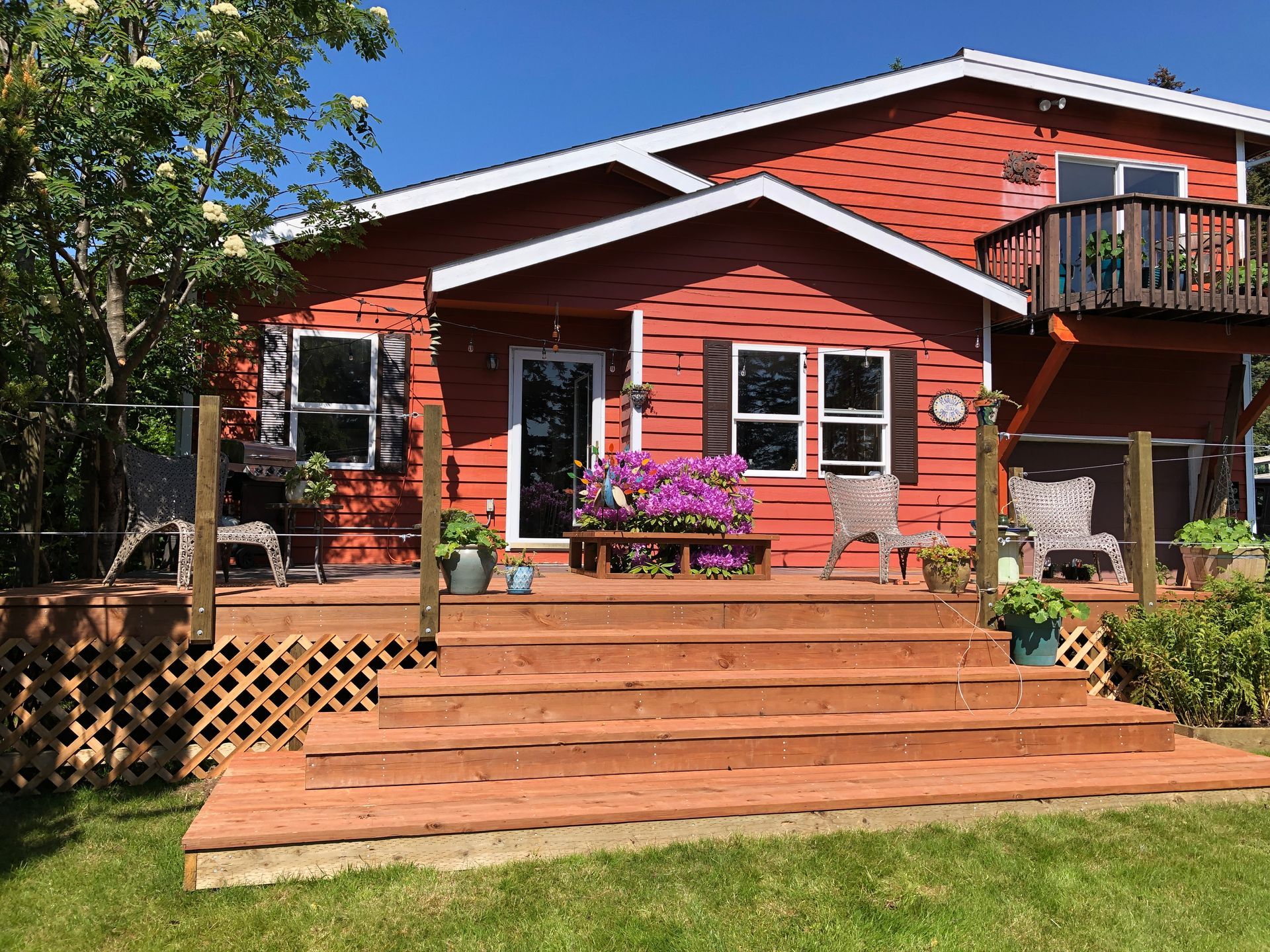 Red house with wooden deck and stairs, potted flowers, and balcony in a sunny yard.