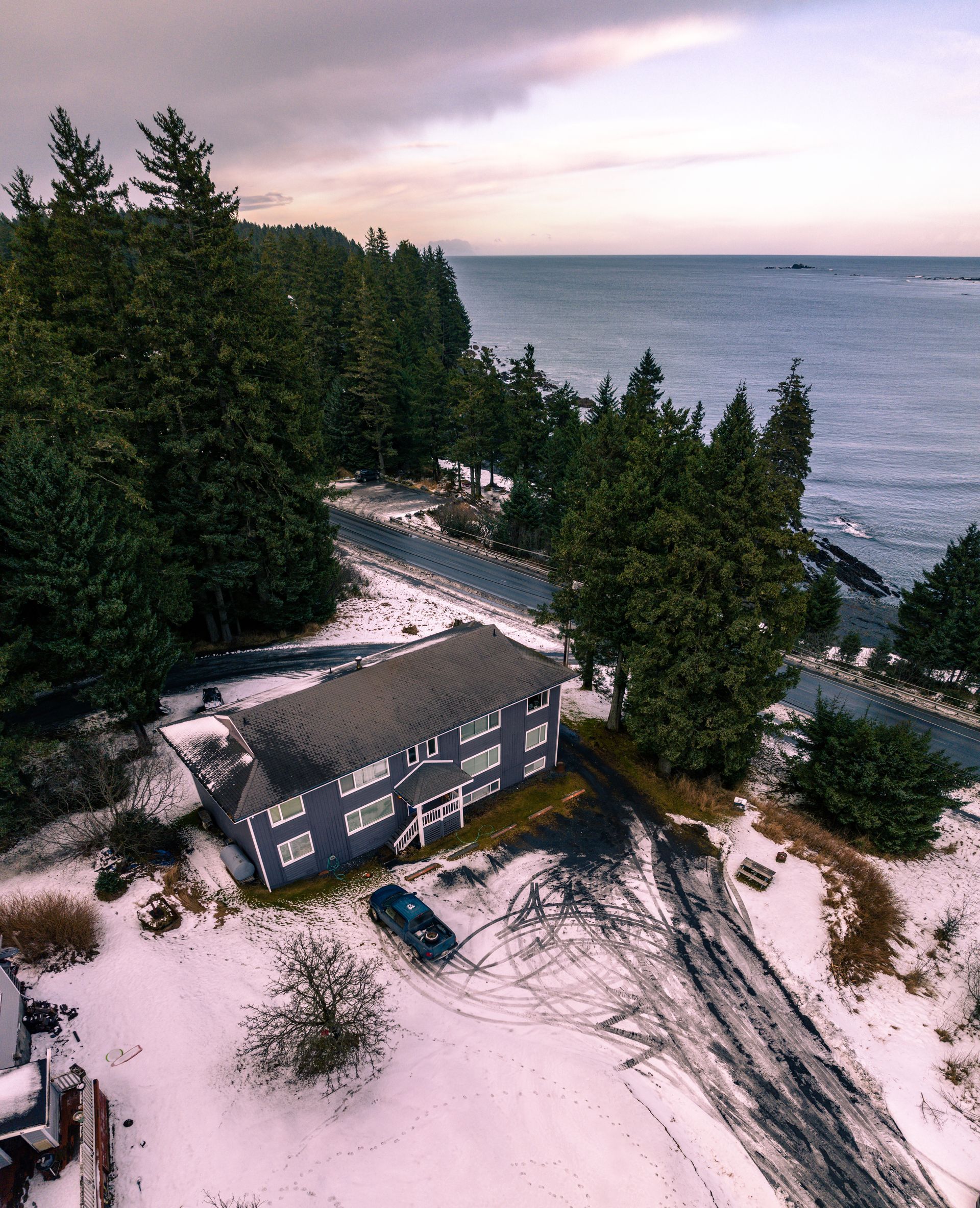 Snowy coastal house beside a forest and ocean at dusk