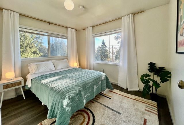 Bright bedroom with a bed, large windows, soft lamps, and a rug in a sunny corner.