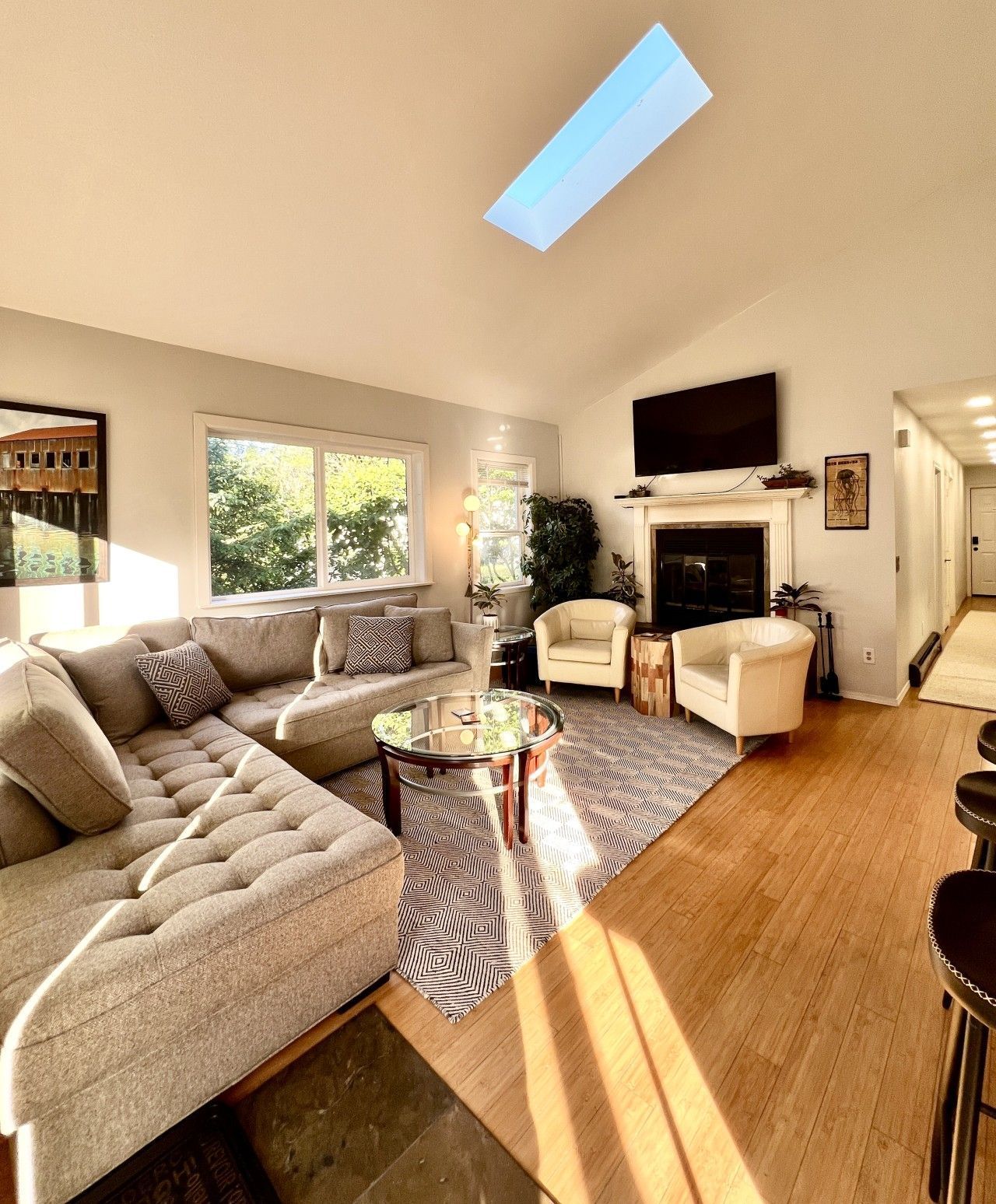 Bright living room with gray sectional, glass coffee table, fireplace, wall TV, and sunlight on wood floor