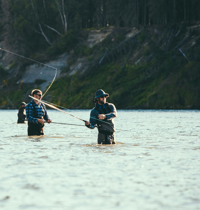 Two anglers wade in a river, casting fishing lines with a forested bank behind them.