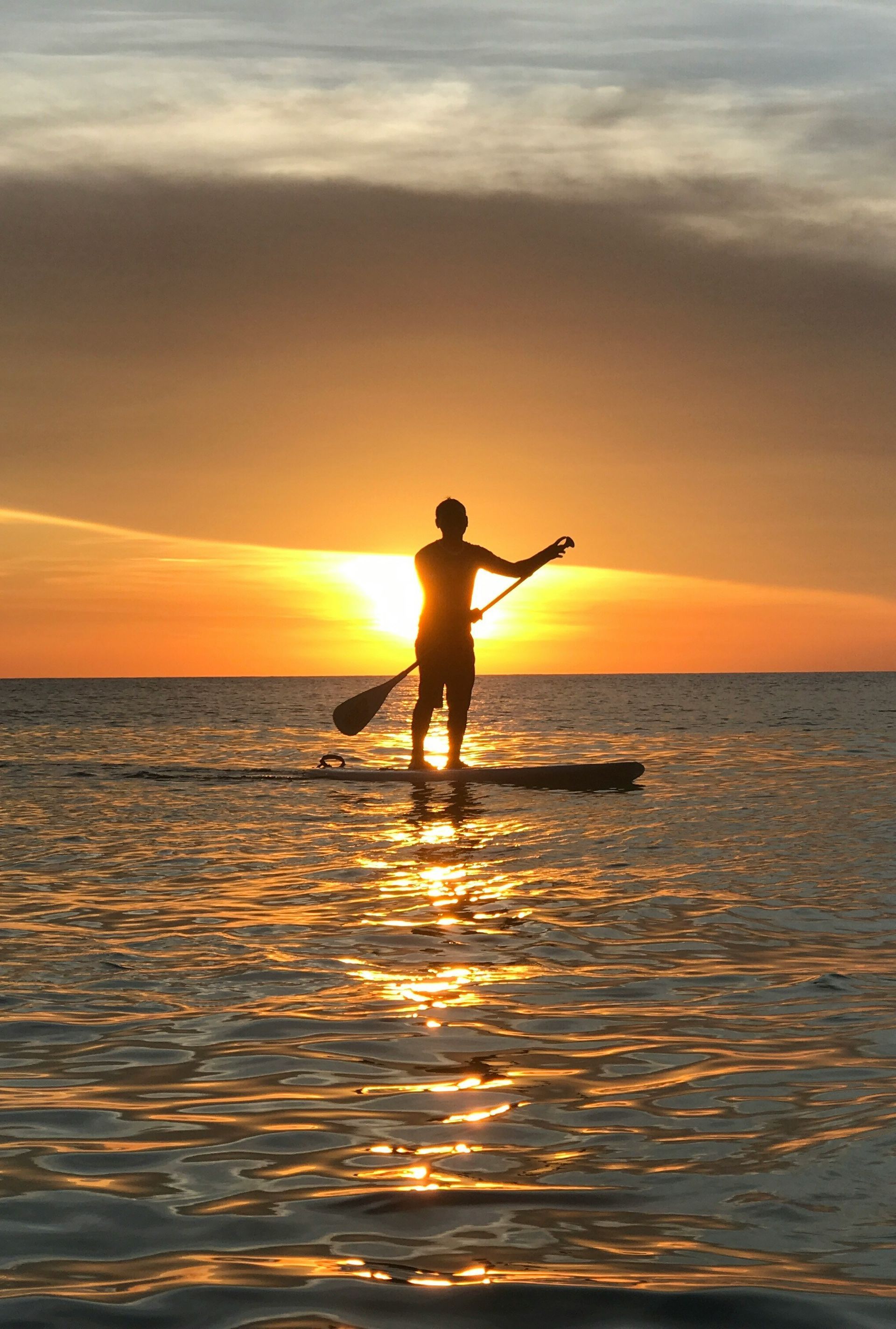 Silhouette of a person paddleboarding at sunset on calm water, with orange sky and reflections.