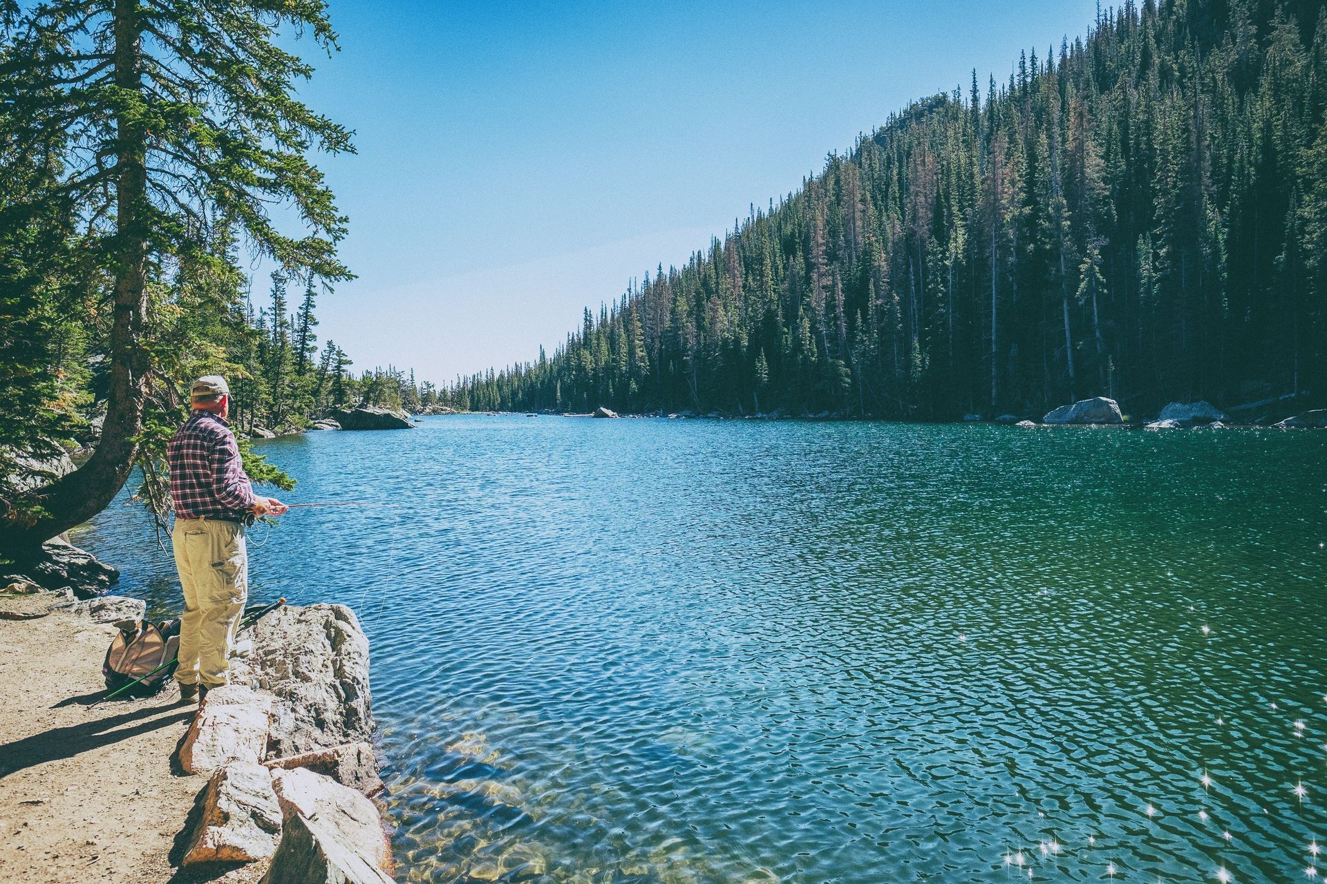 Person fishing beside a clear mountain lake with pine forest and rocky shore