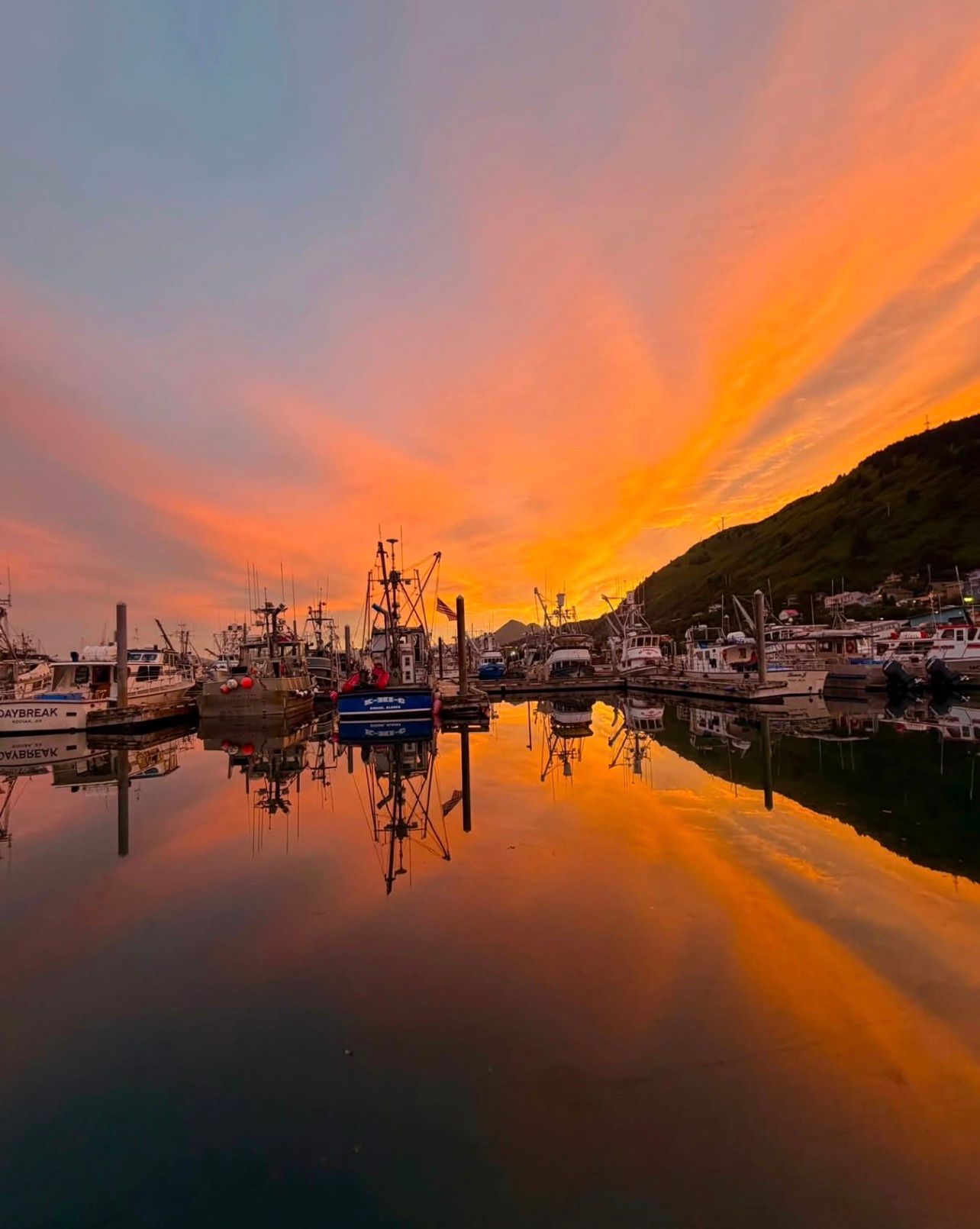 Sunset over a marina with fishing boats, glowing orange sky reflected on calm water