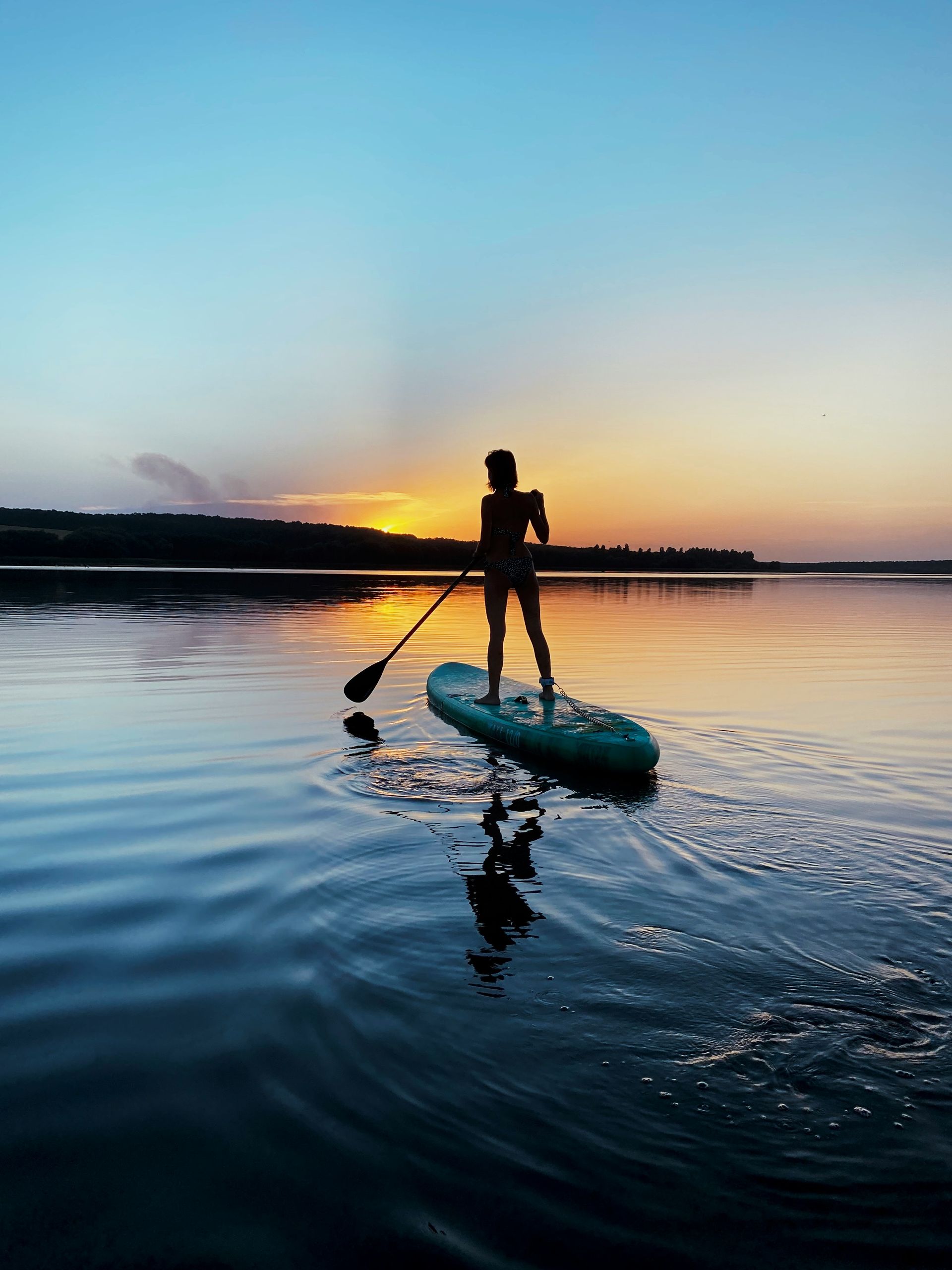 Silhouette paddleboarder on calm water at sunset, with a blue sky and glowing horizon