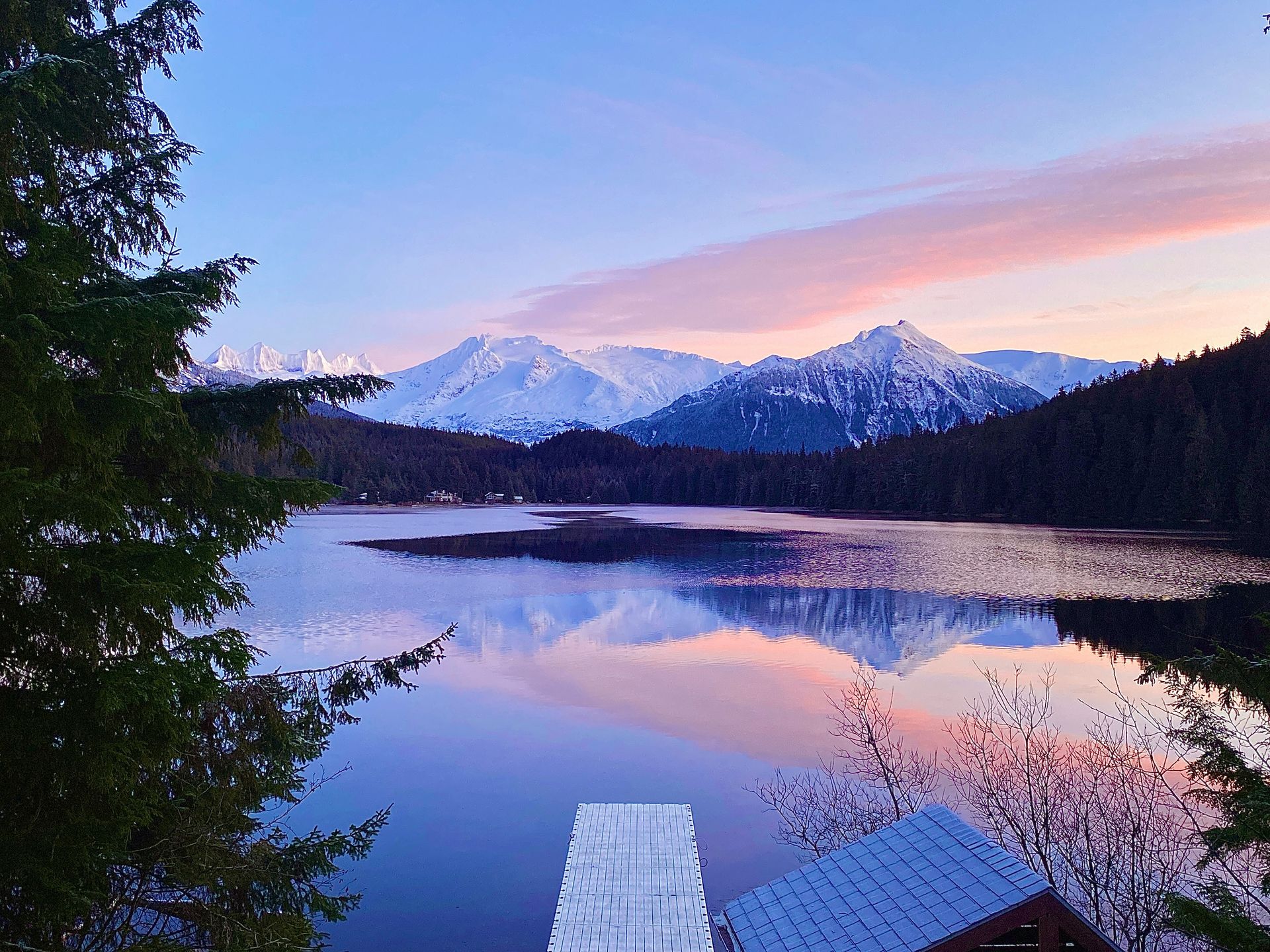 Snowy mountains reflected in a calm lake at sunset, with a dock and dark trees in the foreground