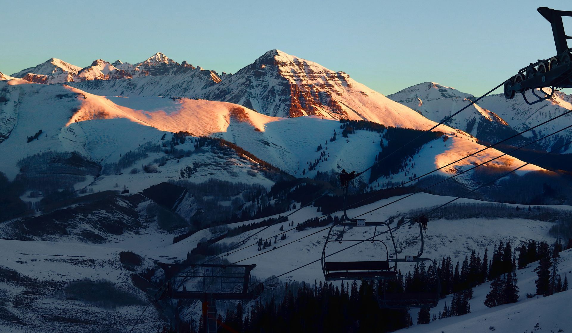 Snow-covered mountain peaks at sunset with pink-orange light and ski lift cables in the foreground