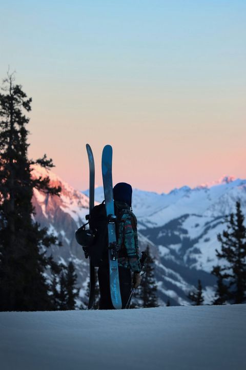 Skis standing in snow at sunset, with mountain peaks and pine trees in the background.