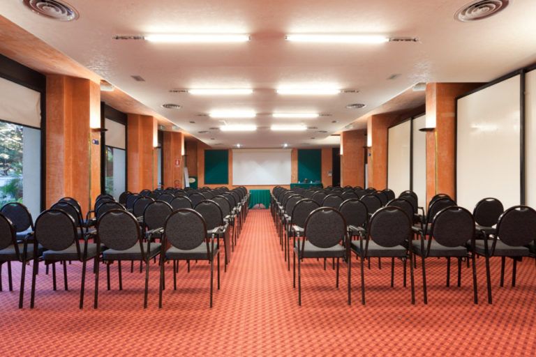 Una sala conferenze con file di sedie nere di fronte a uno schermo di proiezione, caratterizzata da moquette e colonne arancioni.