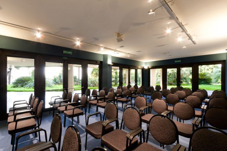 Una sala conferenze con file di sedie marroni di fronte a una serie di grandi porte a vetri che si affacciano su un giardino.
