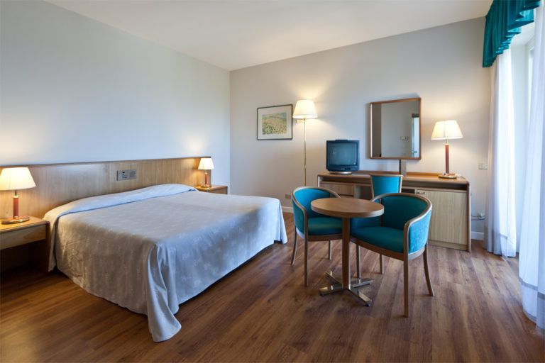 Una camera d'albergo con un letto ben rifatto, un tavolino rotondo con due sedie color verde acqua, una TV e pavimento in legno.