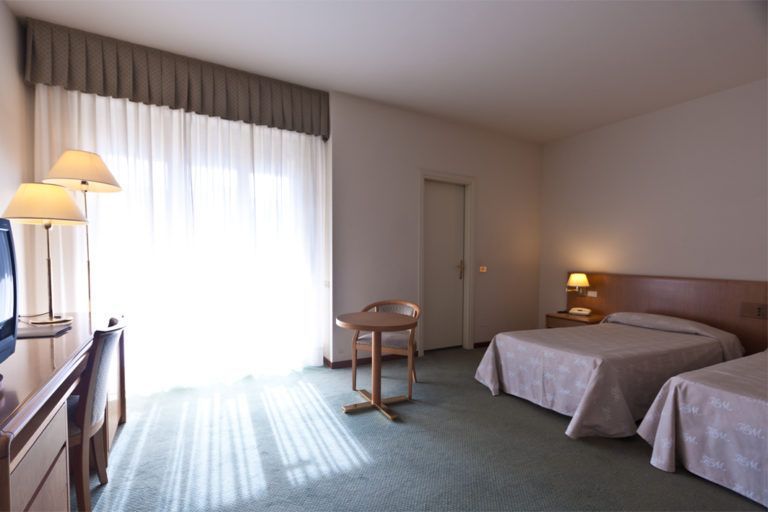 Una camera d'albergo con due letti già fatti, un tavolo con sedia e una scrivania, illuminata dalla luce naturale che entra da una finestra.