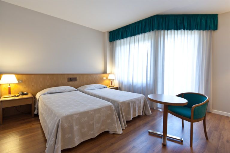 Una camera d'albergo con due letti singoli, un comodino in legno con una lampada e un tavolino in legno con una sedia color verde acqua.