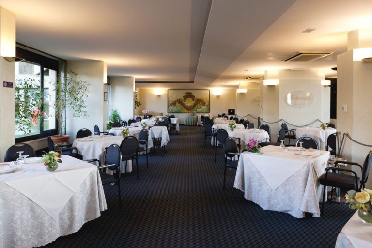 Una sala da pranzo con file di tavoli con tovaglie bianche, sedie nere e un tappeto scuro a motivi geometrici.