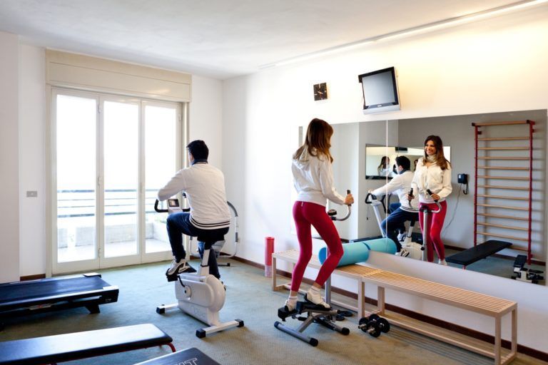 Due persone si allenano su attrezzi da palestra, una cyclette e uno stepper, all'interno di una stanza luminosa con un grande specchio a parete.