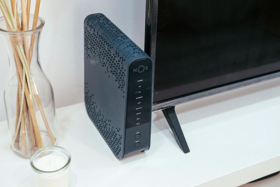 Un router internet nero è appoggiato accanto a uno schermo televisivo su una superficie bianca, con un vaso di canne e una candela nelle vicinanze.