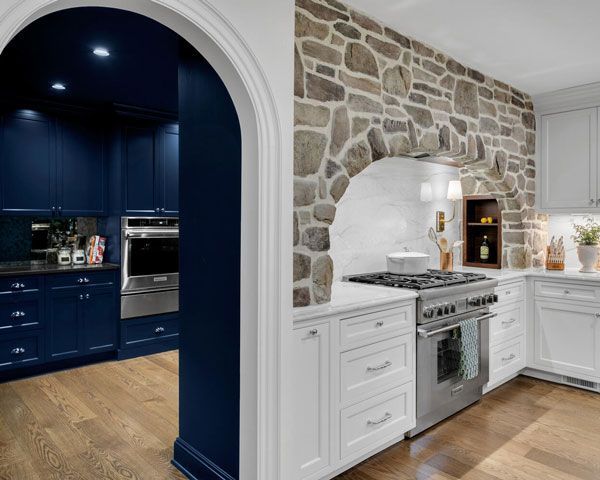 Split kitchen with blue cabinetry on one side and white cabinets, stone backsplash, and stainless stove on the other.