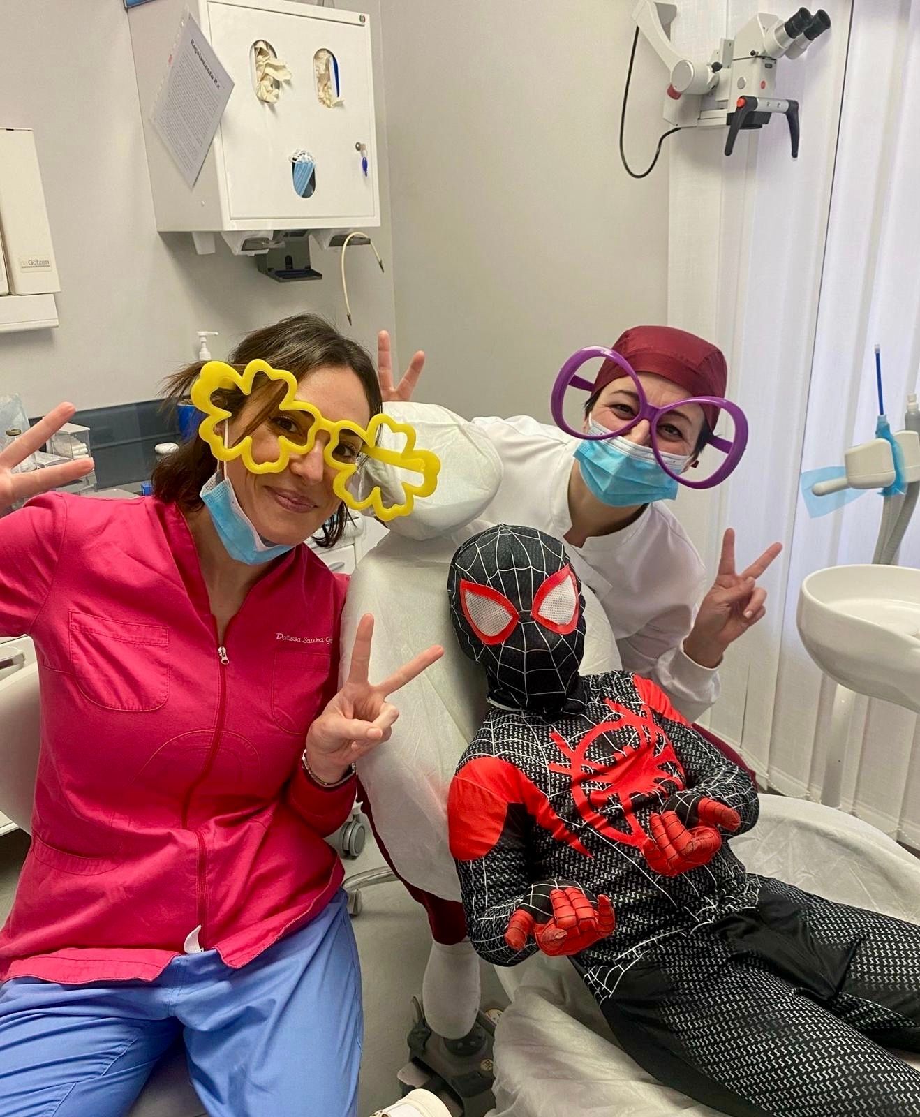 Un bambino in costume da Spider-Man con due dentisti in uno studio dentistico, tutti con occhiali stravaganti, che fanno il segno della pace.