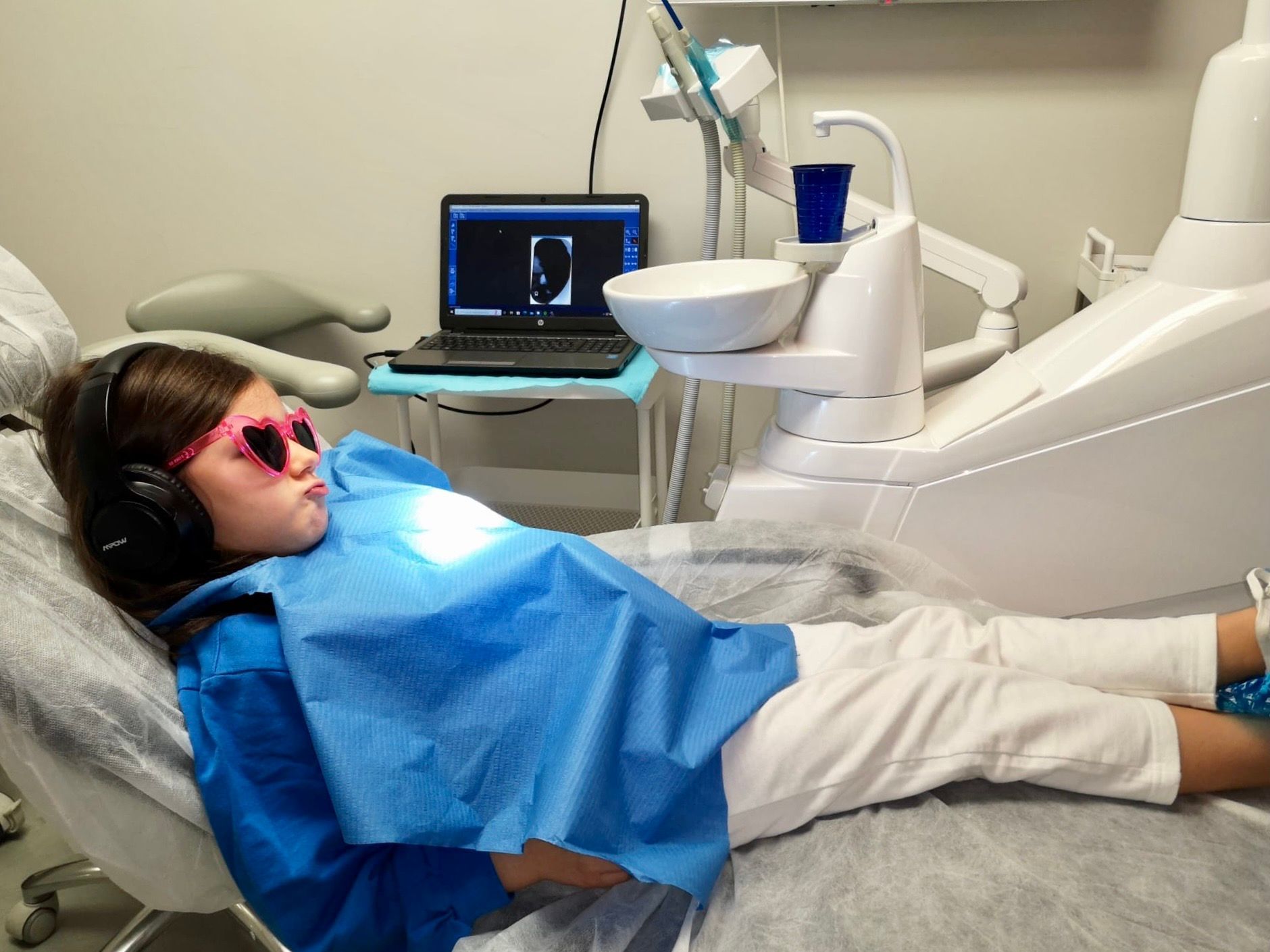 Bambino sulla poltrona del dentista con cuffie, occhiali da sole a forma di cuore rosa e mantello blu, che guarda altrove.