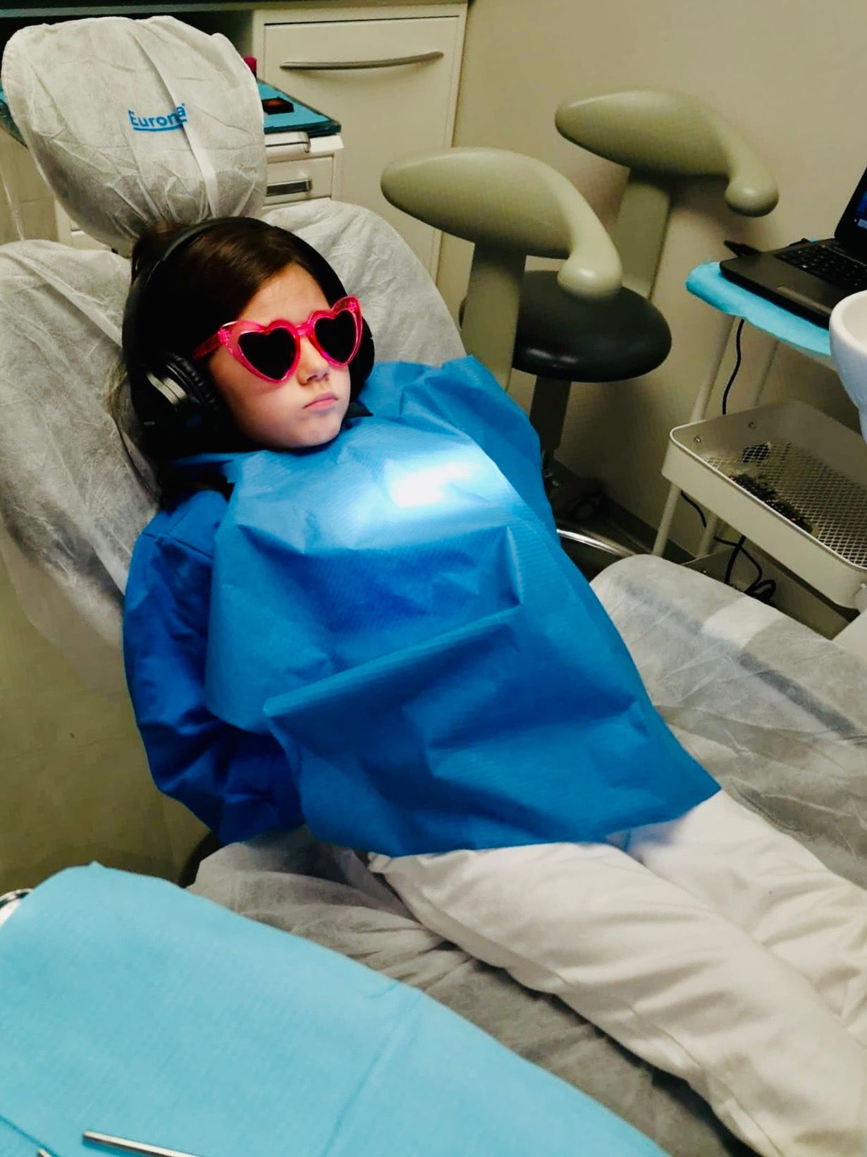 Bambino sulla poltrona del dentista con occhiali a forma di cuore e cuffie, coperto da un bavaglino blu, sotto una luce intensa.