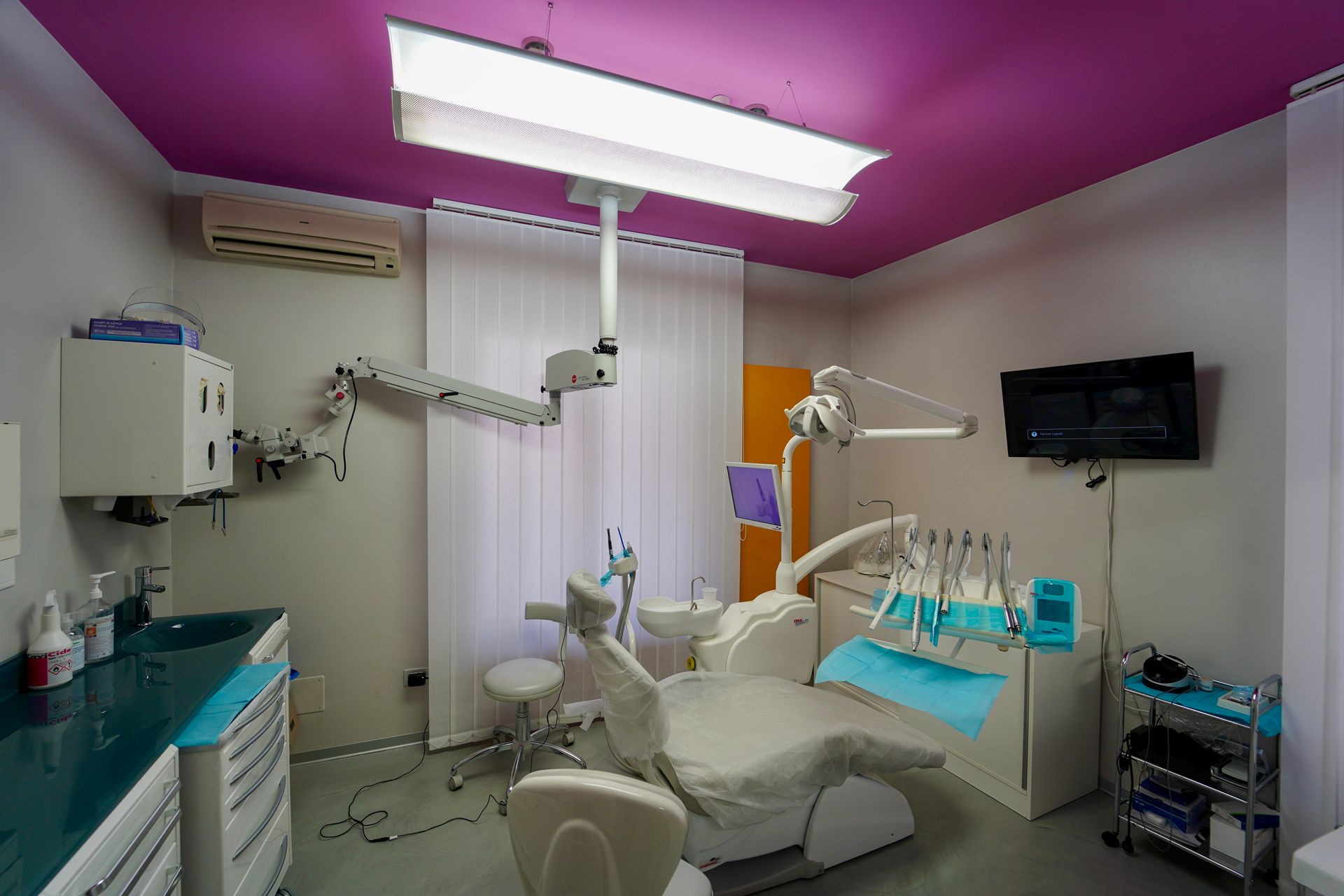 Interno dello studio dentistico con soffitto rosa, poltrona odontoiatrica, attrezzatura e una piccola TV.