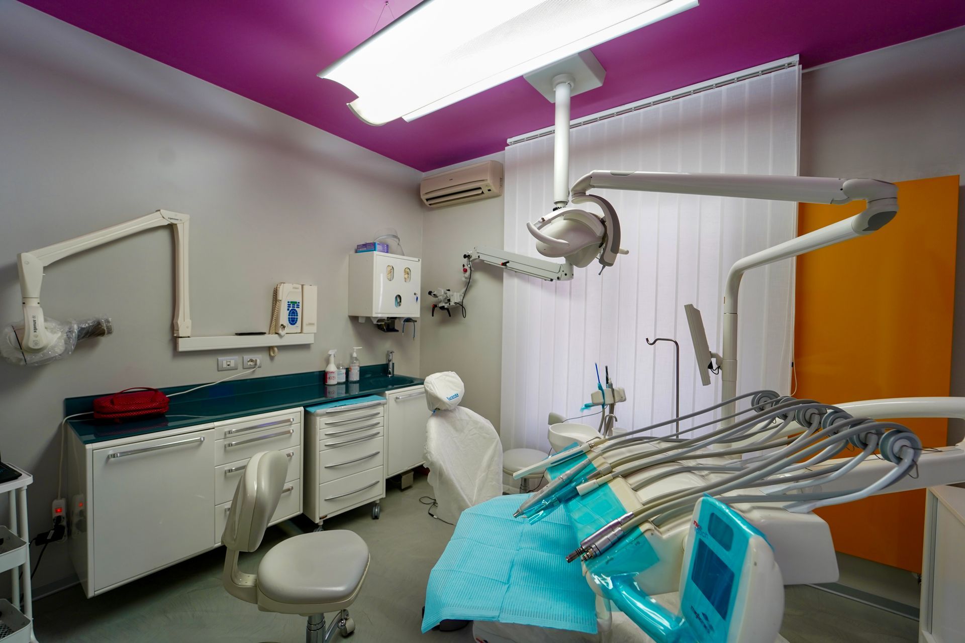 Sala visita odontoiatrica con poltrona, attrezzatura e armadietti. Soffitto viola, pareti bianche e dettagli gialli.