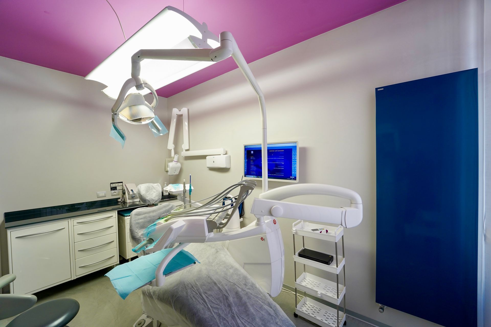 Sala visita odontoiatrica con poltrona, luce e attrezzatura; soffitto rosa, dettagli blu e bianchi.