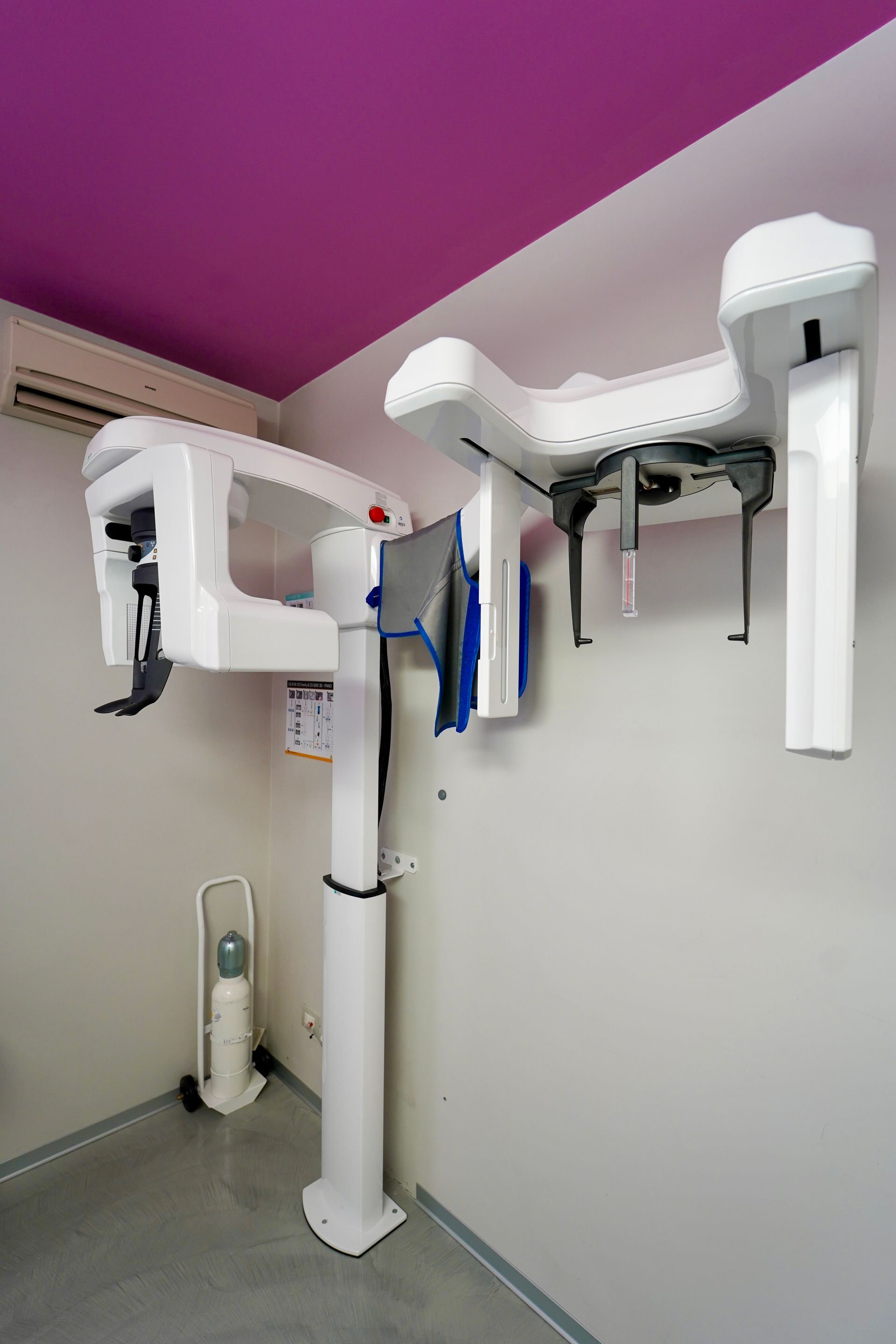 Apparecchio radiografico panoramico dentale montato su una parete bianca. La stanza ha un soffitto viola e un pavimento grigio.
