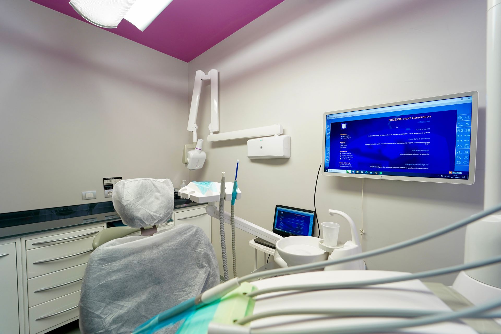 Una sala per visite odontoiatriche con una sedia per visite, strumenti odontoiatrici e uno schermo per computer montato sulla parete.