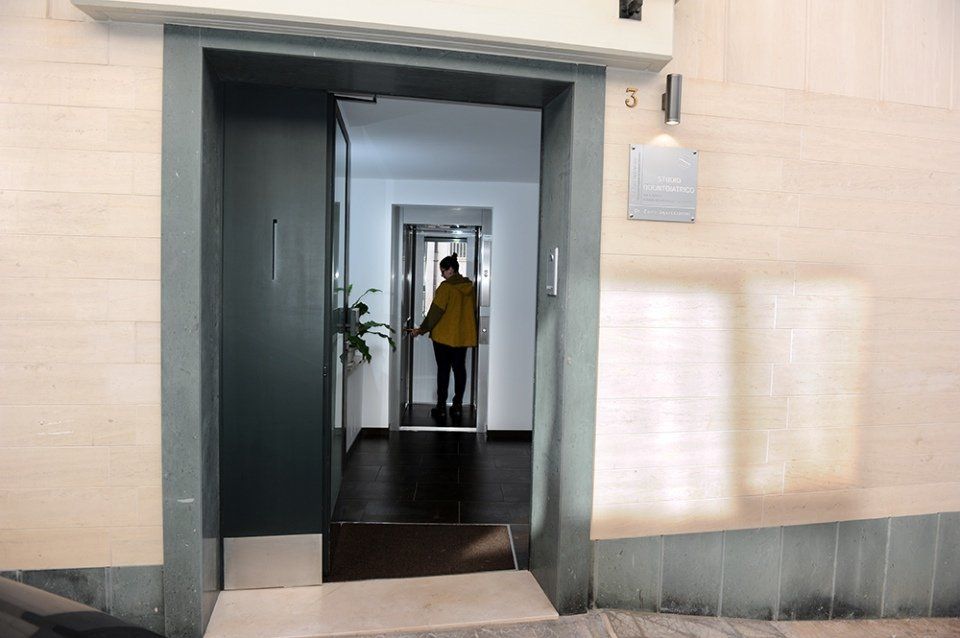 Una persona con la giacca gialla entra in un ascensore. L'ascensore si trova nel corridoio di un edificio, con una parete bianca simile a mattoni.