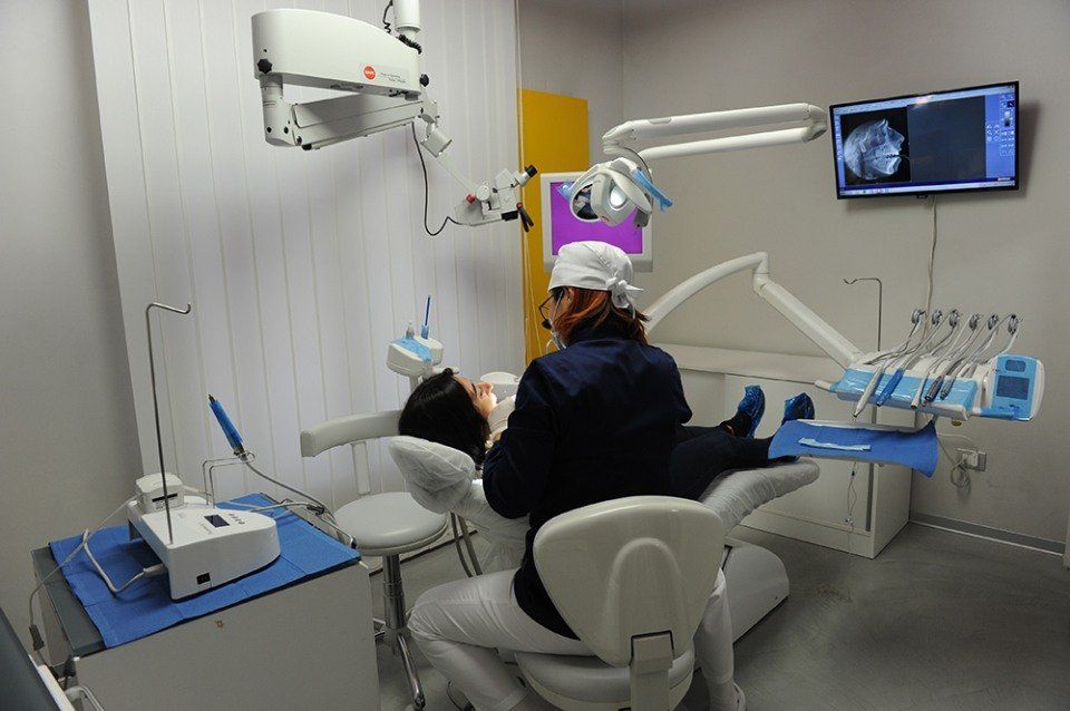 Dentista che visita un paziente su una poltrona odontoiatrica; apparecchiature mediche visibili, monitor che mostra una radiografia.