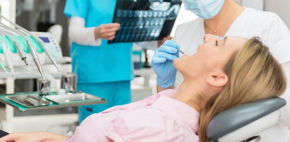 Un dentista esamina i denti di un paziente. Il paziente è sdraiato su una poltrona, con la bocca aperta. Un'assistente dentale tiene in mano le radiografie.