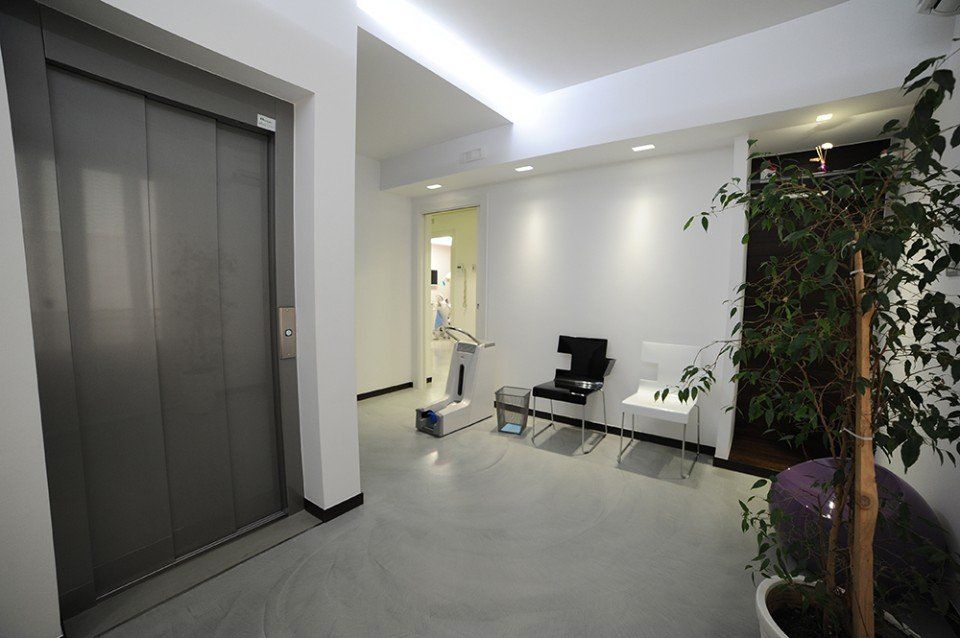 Sala d'attesa con ascensore, attrezzature mediche, sedie e pianta in vaso. Interni luminosi e moderni con pavimento grigio.