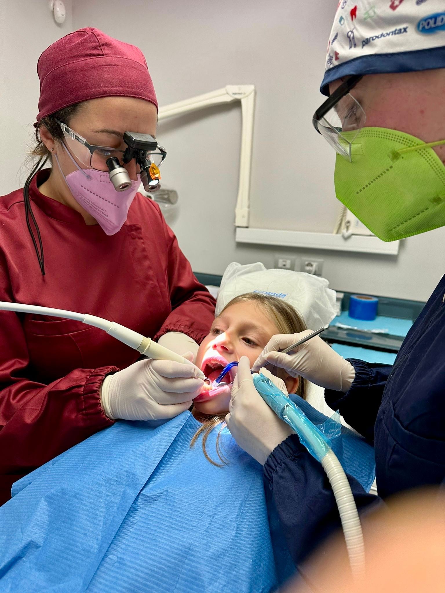 Dentista che cura i denti di un bambino. Dentista che indossa mascherina, lenti d'ingrandimento e guanti, tiene gli strumenti in bocca. È presente un assistente.