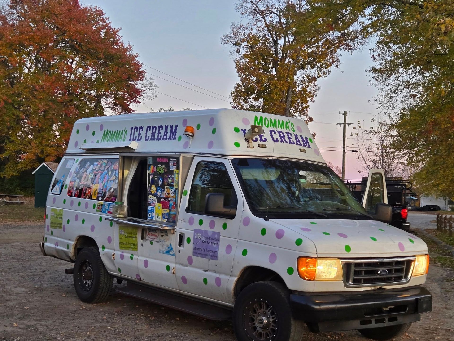 White ice cream truck with polka dots and colorful menu graphics, parked on a gravel lot near trees with autumn leaves.