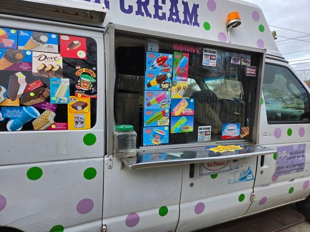 A white ice cream truck with green and purple polka dots, featuring a service window display of various ice cream treats.